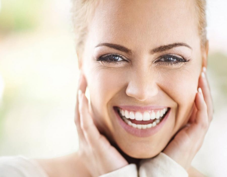 Closeup portrait of an attractive mid 30's woman gently touching her neck and smiling while looking at the camera and sincerely smiling. Blurry green background.